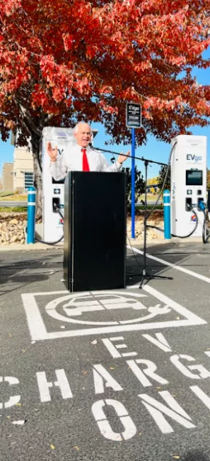 CEO executive director Will Toor speaking at podium in front of DCFC Plazas charger in front of fall red tree