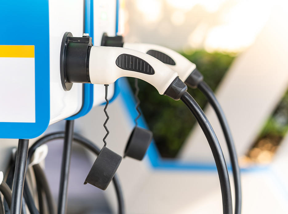 Close-up of an electric vehicle charging station plug in a holster with a blue and white color scheme, indicating a clean energy source. The focus is on the handle of the plug, which is white with black accents and a textured grip, against a softly blurred background.