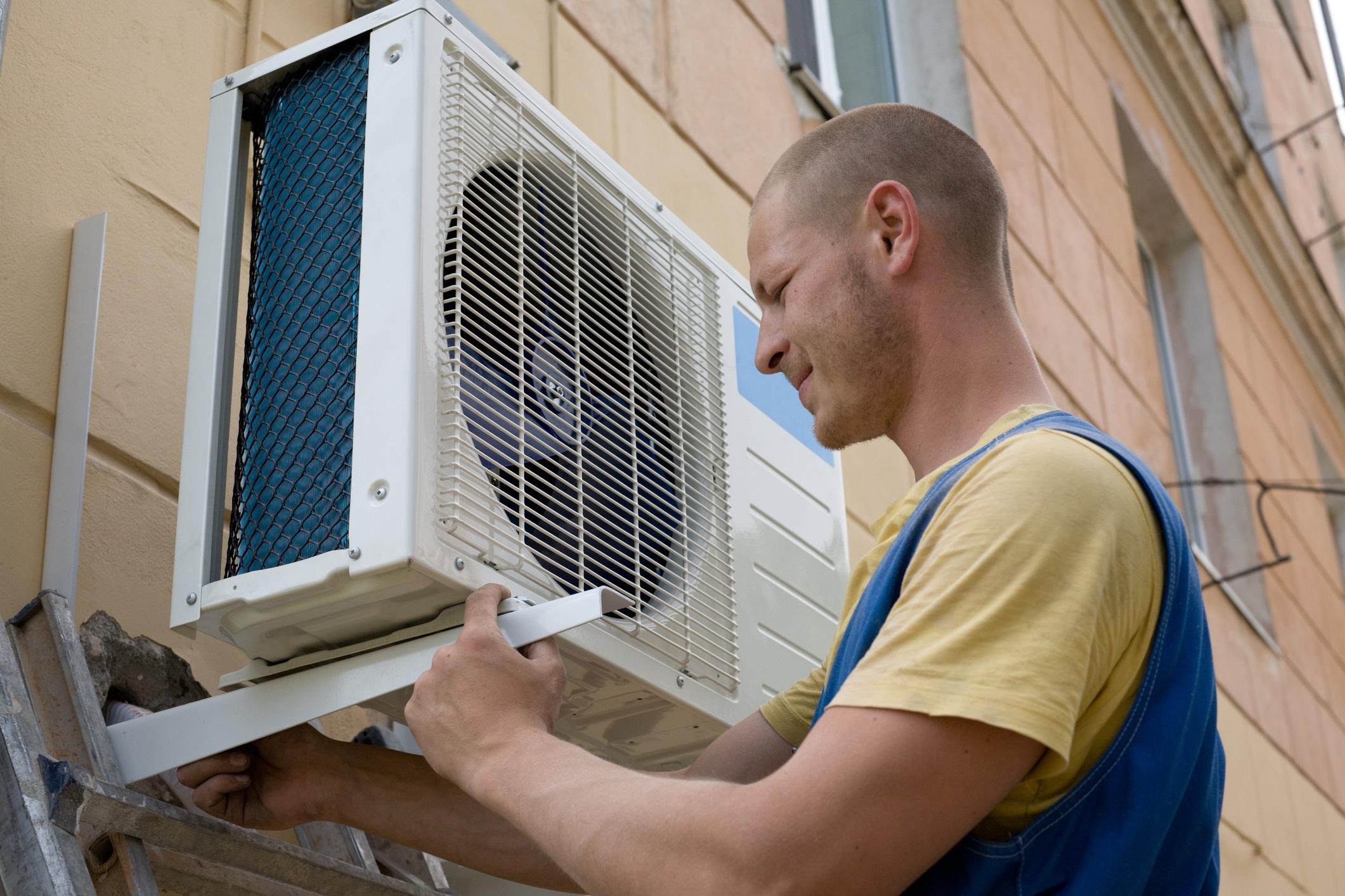 A person installs an air-source heat pump on the side of a multi-story building.