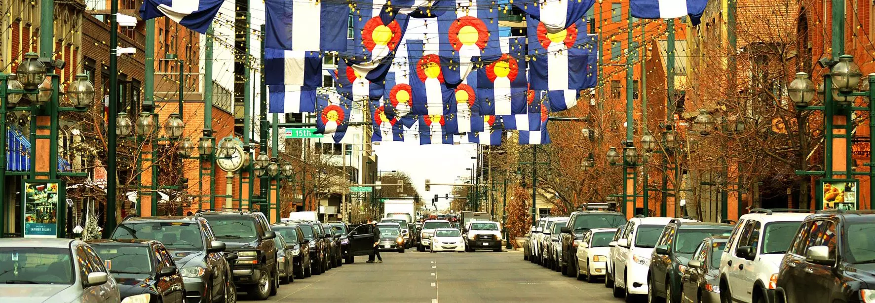 A photograph from the middle of the street facing cars that are stopped at a light in the distance. Cars are parked on both sides of the street, and many Colorado flags are hanging over the road from a rope between buildings on each side.