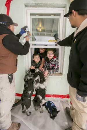 Three children crowd a window from inside a house watching two workmen caulk seal the exterior. Three dogs on the outside are excited to see the children by the window.