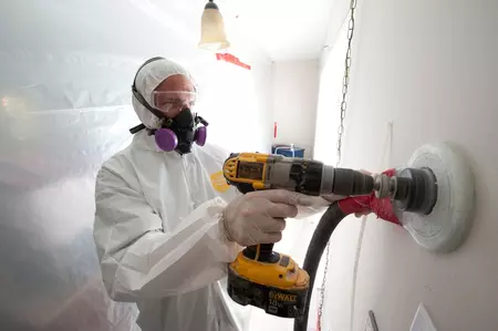 A man wearing a respirator and white coveralls drilling a hole into a wall