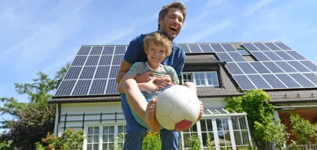 father holding son in front of home with rooftop solar under a clear blue sky