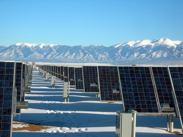 A large solar panel array stands in a flat, snow-covered landscape under a clear blue sky. The photo is taken from a low angle, looking across multiple rows of dark blue solar panels. They are organized in neat, parallel lines that recede into the distance. In the foreground, a light dusting of snow covers the ground, with some footprints visible. In the background, a majestic mountain range dominates the horizon, their snow-covered peaks jagged and distinct against the sky. The image juxtaposes the human-m