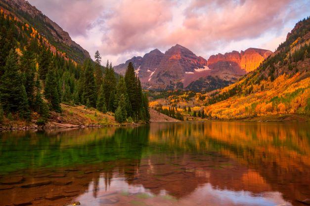 A photograph of a mountain (maroon bells) in the background with a lake in the foreground at sunset. The mountain is reflected in the water. The picture was taken autumn, with some leaves showing bright yellow coloring.